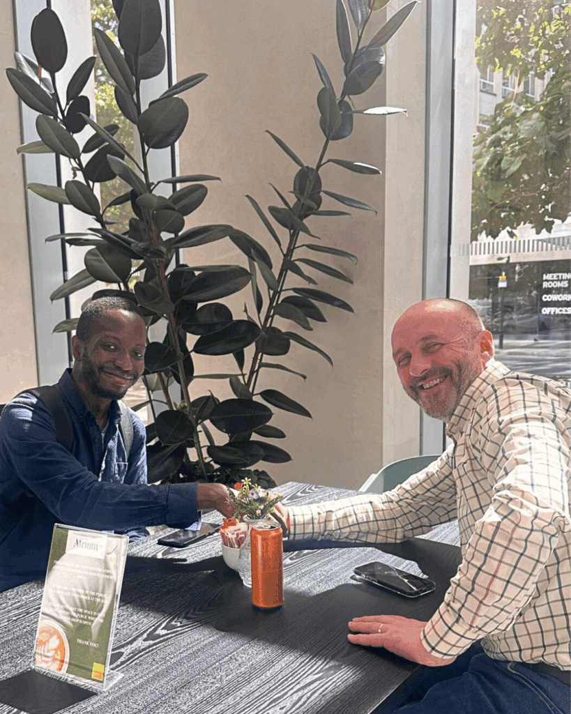 Two men smiling and shaking hands across a café table, with a soft drink and potted plant in the background, conveying a positive meeting.