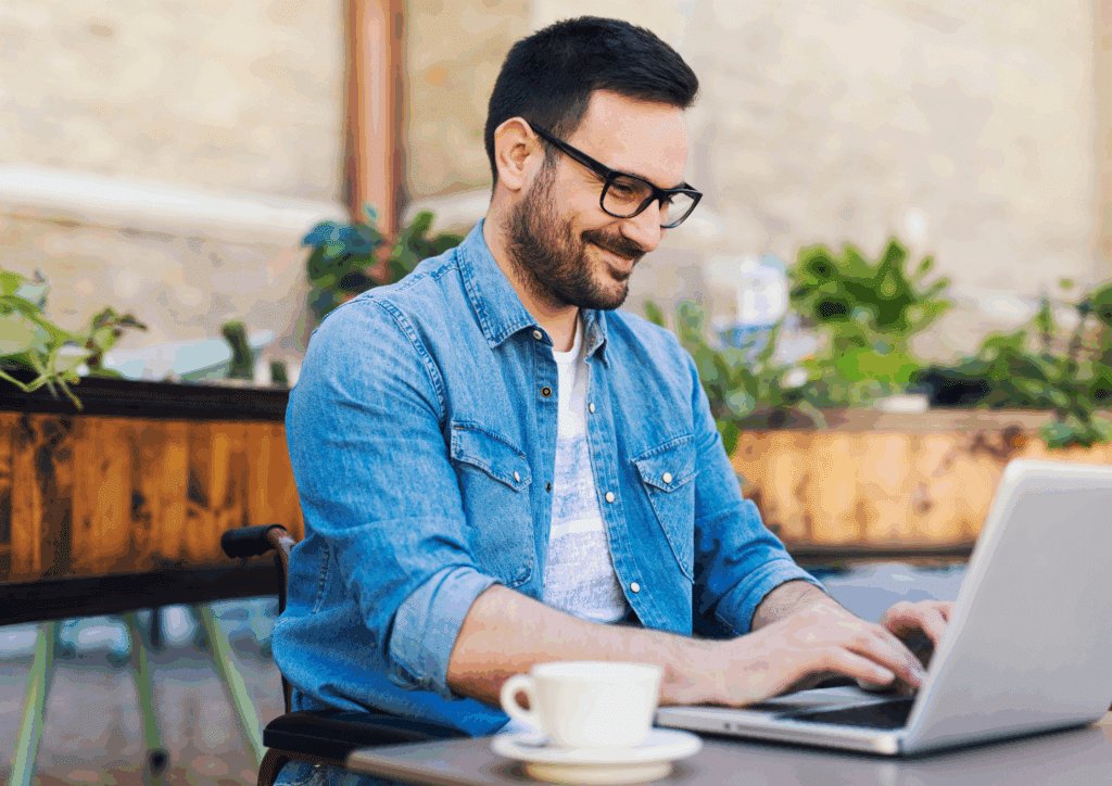A smiling man in glasses and a denim shirt types on a laptop at an outdoor café table, with a cup of coffee and potted plants in the background.