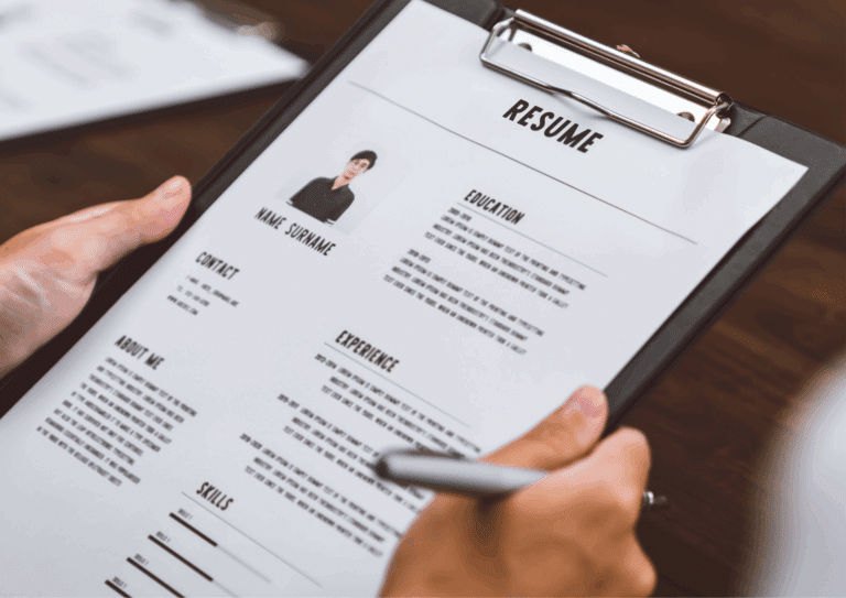 Close-up of a hand holding a resume on a clipboard with a pen, preparing to make notes. The CV includes a profile photo, contact information, experience, education, and skills.