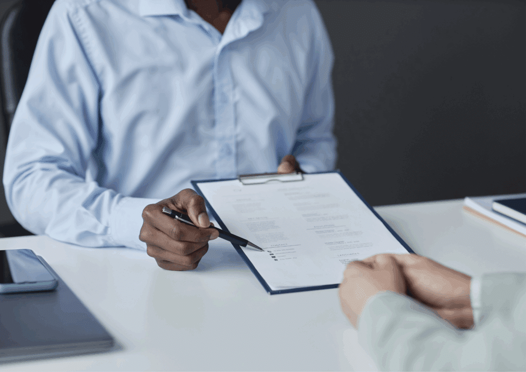 A recruiter or hiring manager in a blue shirt holding and reviewing a candidate’s CV across a desk during an interview or recruitment meeting.