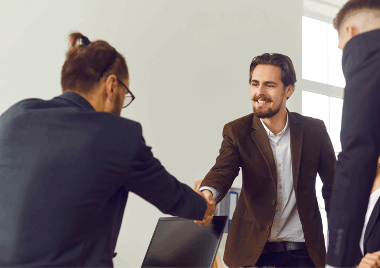 A smiling man in a brown blazer shaking hands with another person during a professional meeting or job interview.