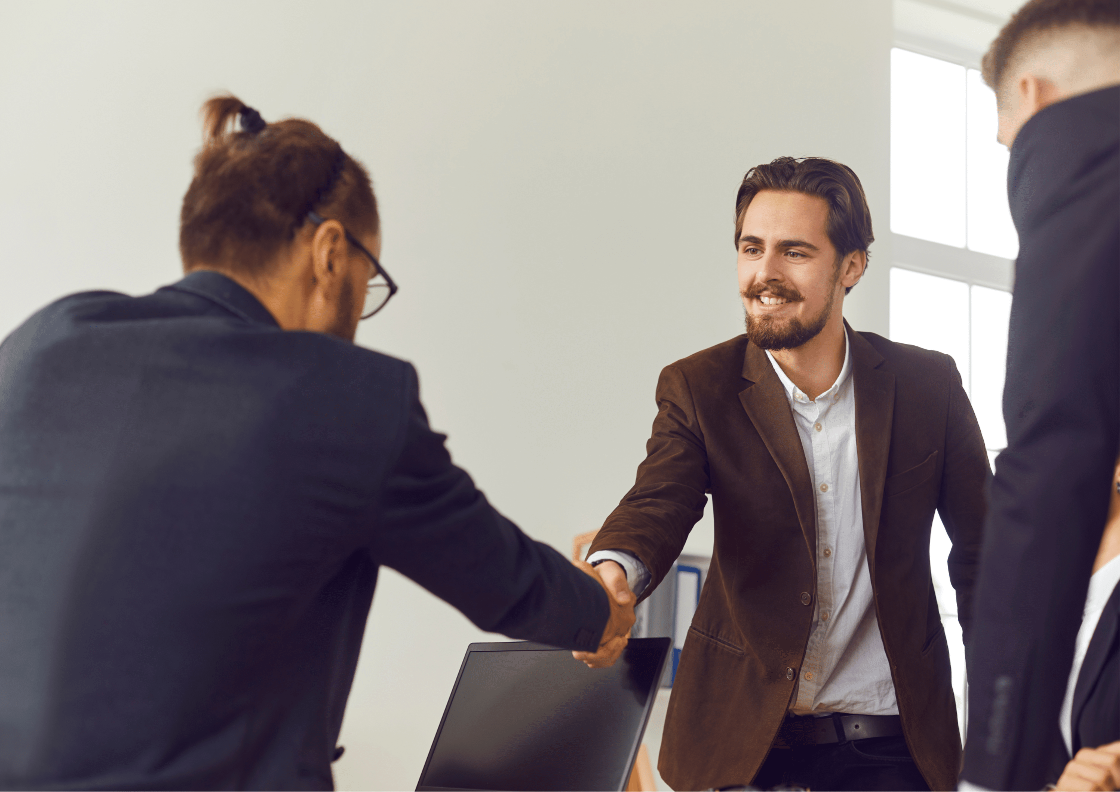 A smiling man in a brown blazer shaking hands with another person during a professional meeting or job interview.