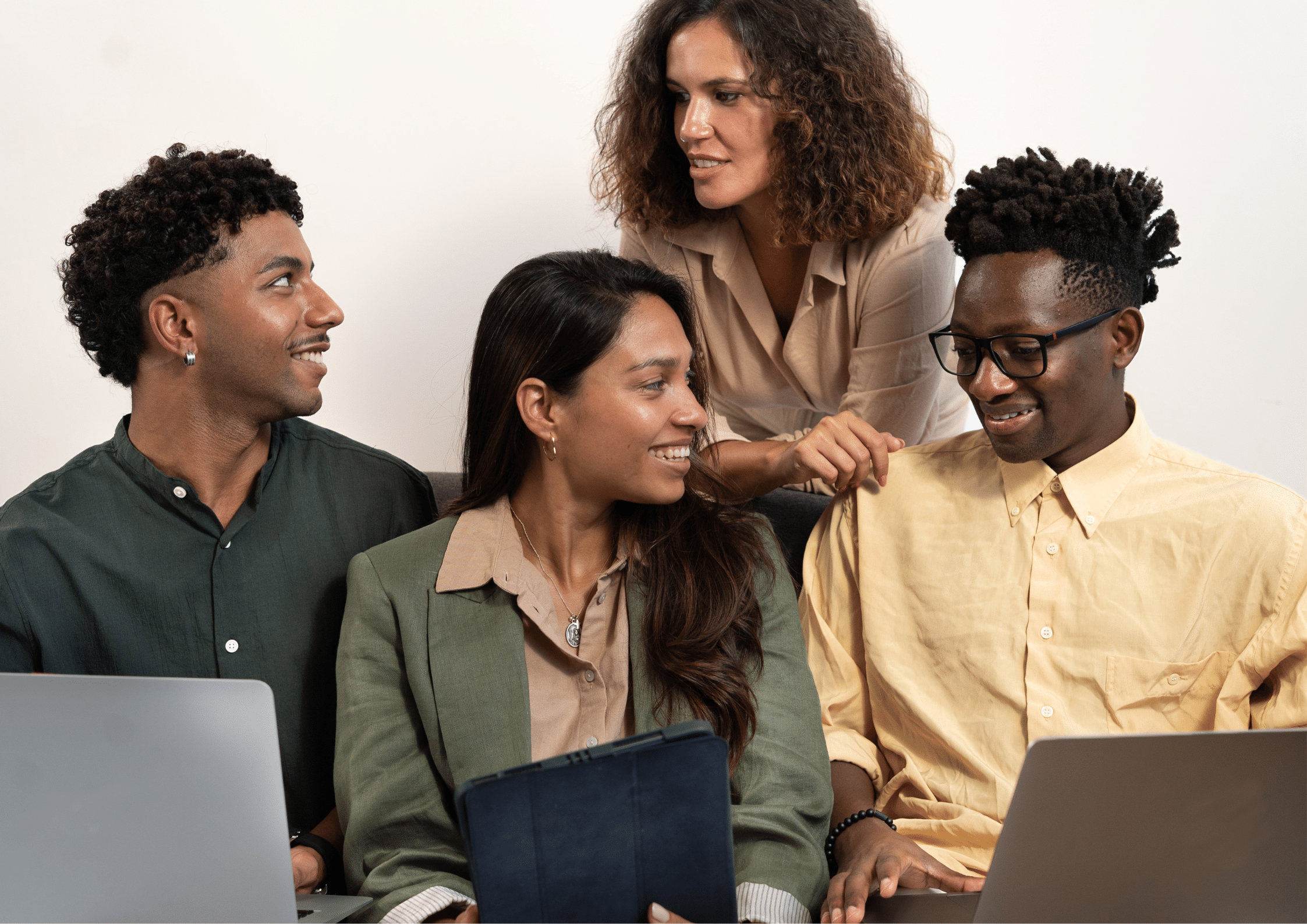 A group of four diverse colleagues smiling and discussing work while using laptops and a tablet