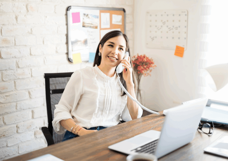 Smiling woman sitting at her desk, speaking on a landline phone while working on a laptop in a modern office.
