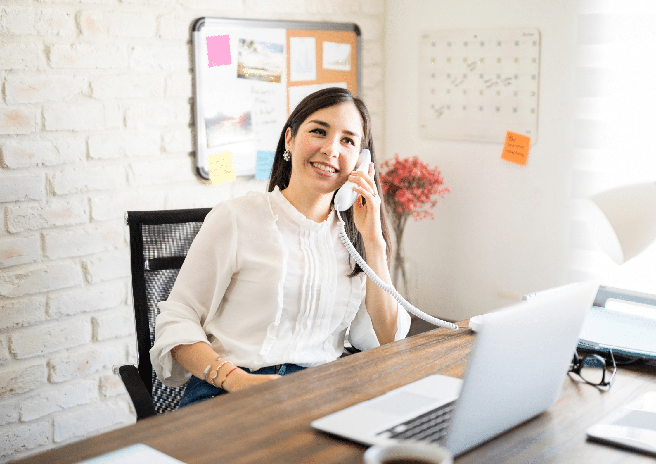 Smiling woman sitting at her desk, speaking on a landline phone while working on a laptop in a modern office.