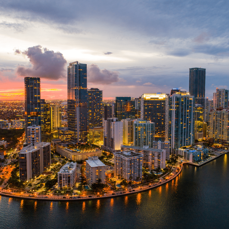 Aerial view of the Miami skyline at sunset, with modern high-rise buildings along the waterfront and warm lights reflecting across Biscayne Bay.