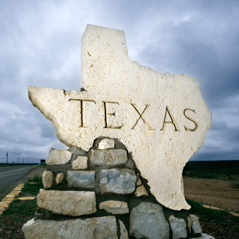 Stone monument shaped like the state of Texas, engraved with the word “TEXAS,” standing beside a quiet highway under a cloudy sky.