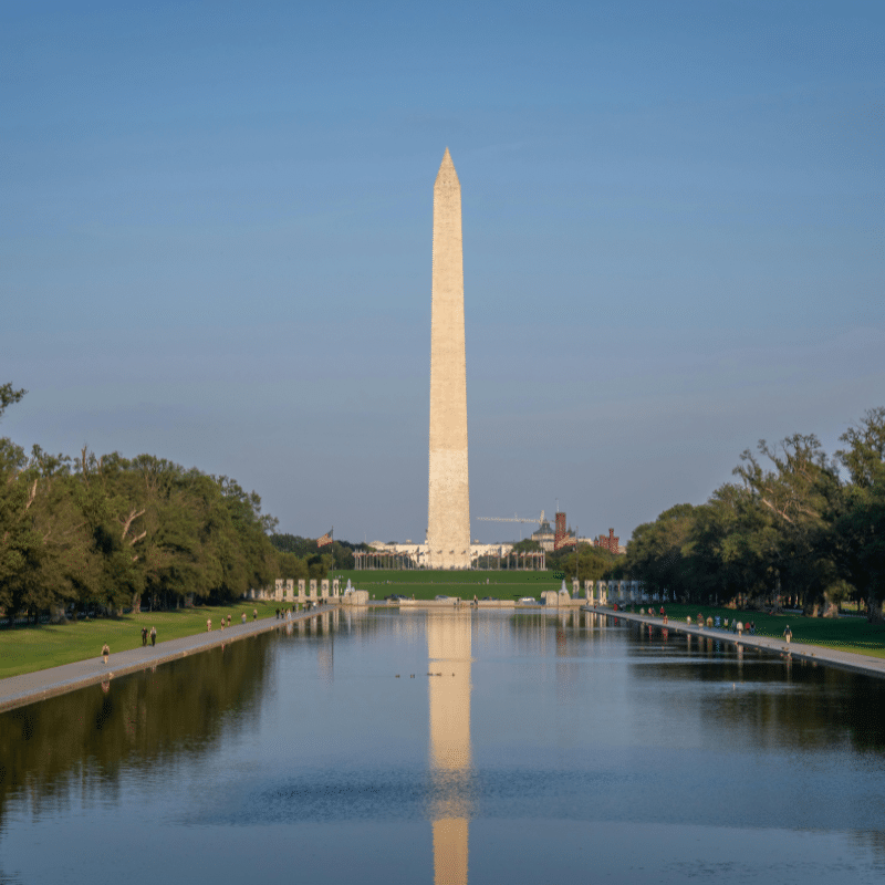 View of the Washington Monument in Washington, D.C., reflected in the calm waters of the Reflecting Pool, surrounded by trees and visitors on a clear day.