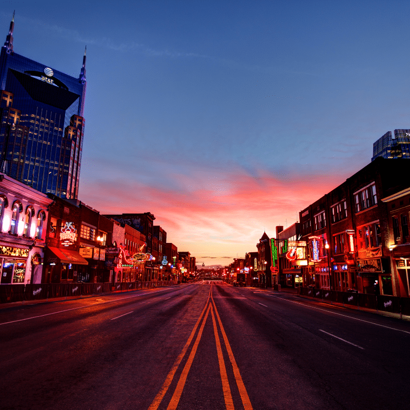 Empty Broadway street in Nashville at sunrise, with colourful neon lights and the AT&T building visible against a vibrant morning sky.