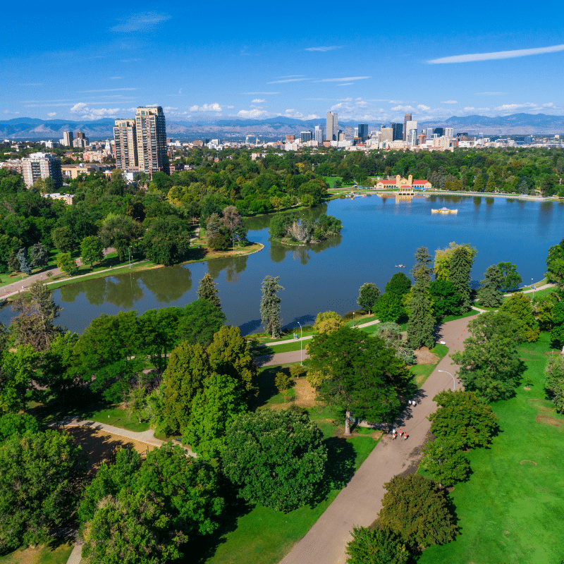 Aerial view of Denver’s City Park with lakes, trees, and the downtown skyline in the distance, set against the Rocky Mountains under a clear blue sky.