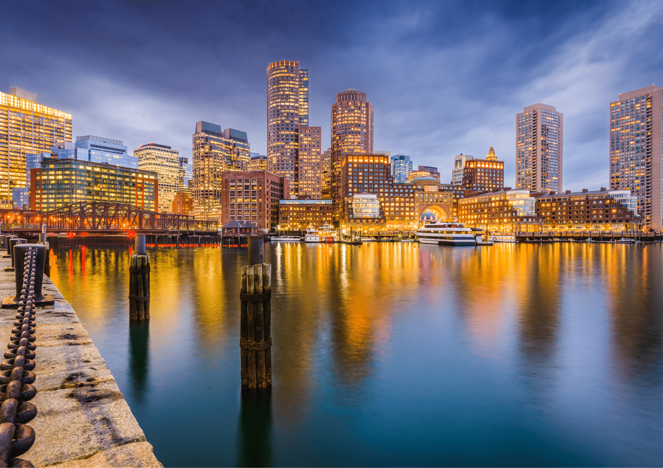 Boston city skyline at dusk with illuminated buildings reflected on the harbor water.