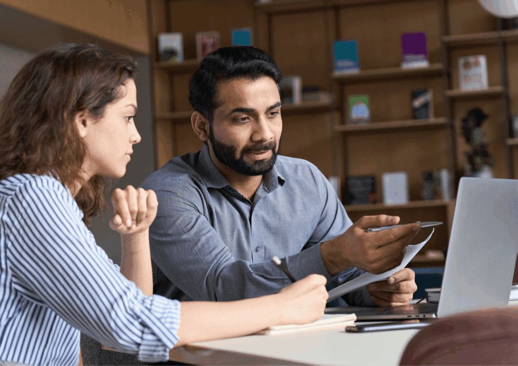 Two professionals sitting at a desk with a laptop, discussing documents and taking notes during a work meeting.