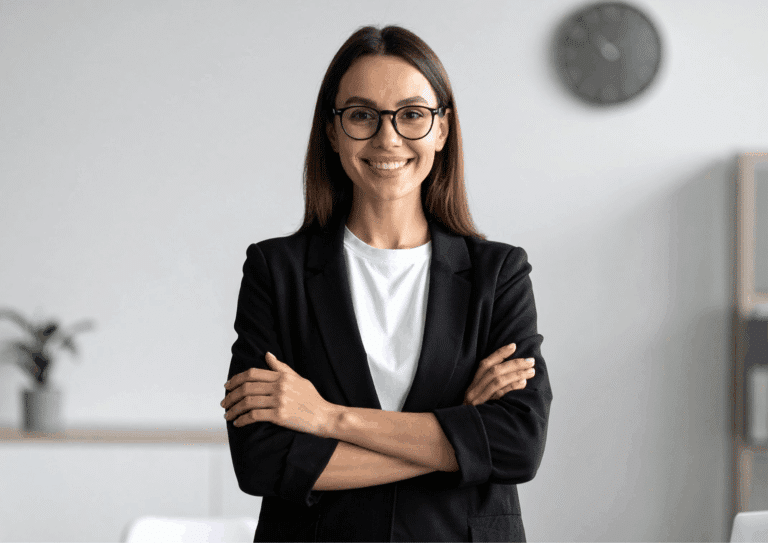 Woman wearing glasses and a black blazer standing with arms crossed, smiling confidently in an office setting.
