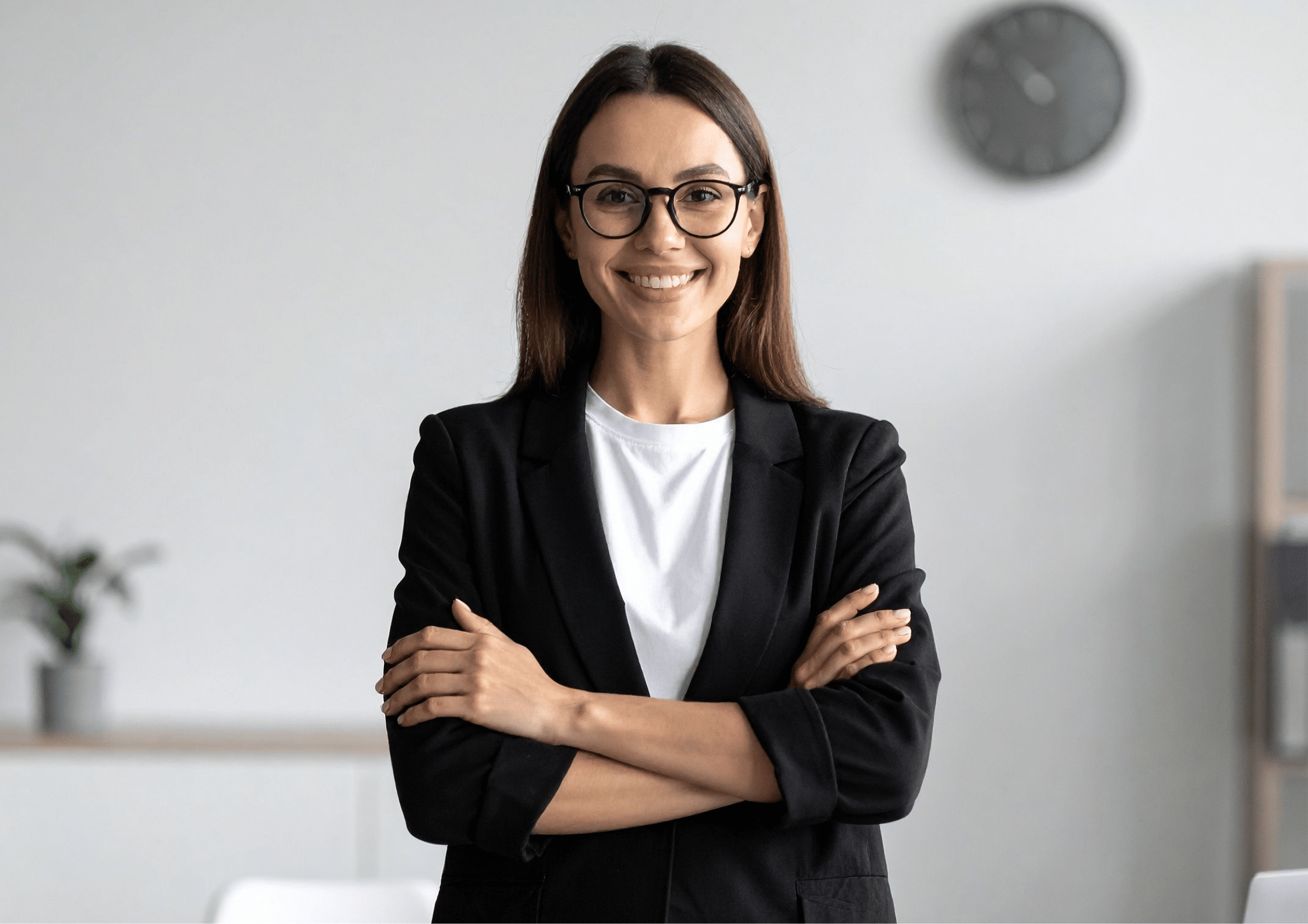 Woman wearing glasses and a black blazer standing with arms crossed, smiling confidently in an office setting.