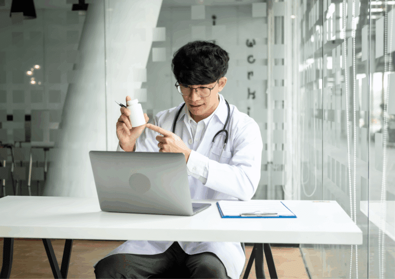 Doctor wearing a white coat and stethoscope holding a medicine bottle and pointing to it during an online video consultation.