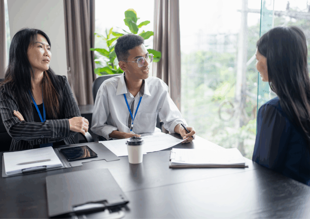 Two hiring managers interviewing a candidate across a desk with documents and coffee cups on the table.