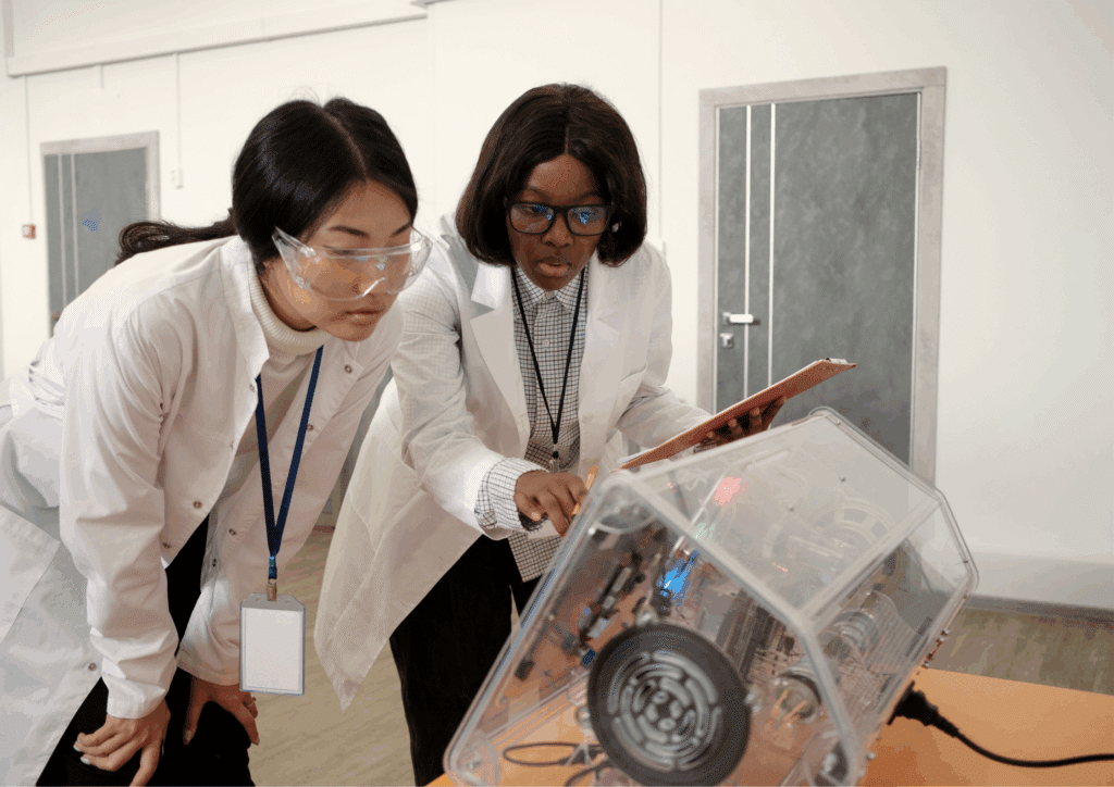 Two researchers in lab coats examining an experimental device, one holding a clipboard and the other taking notes.