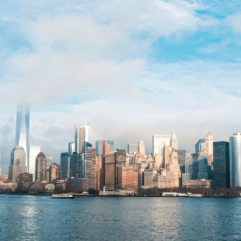 View of the New York City skyline from across the Hudson River, featuring modern skyscrapers under a bright blue sky with scattered clouds.
