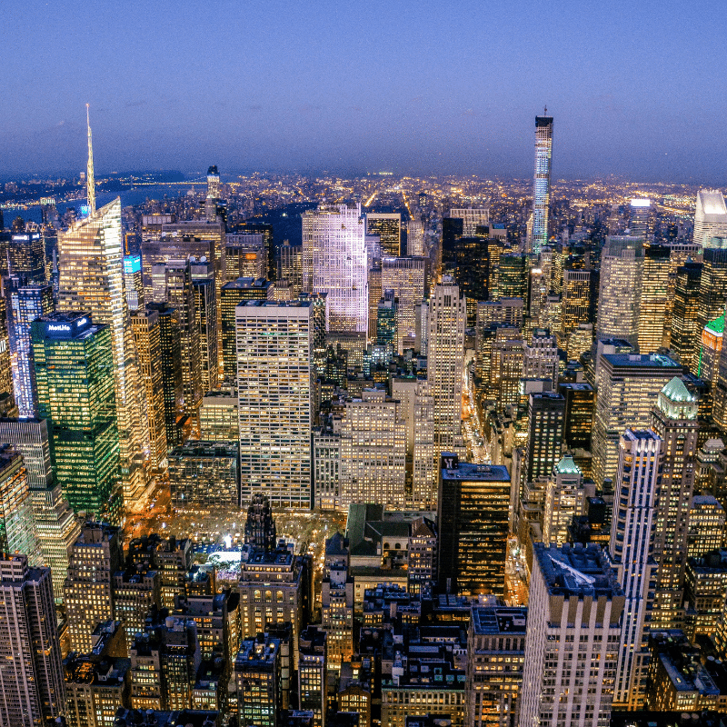 Aerial view of New York City illuminated at night, showing a dense cluster of skyscrapers and bright city lights stretching across the horizon.