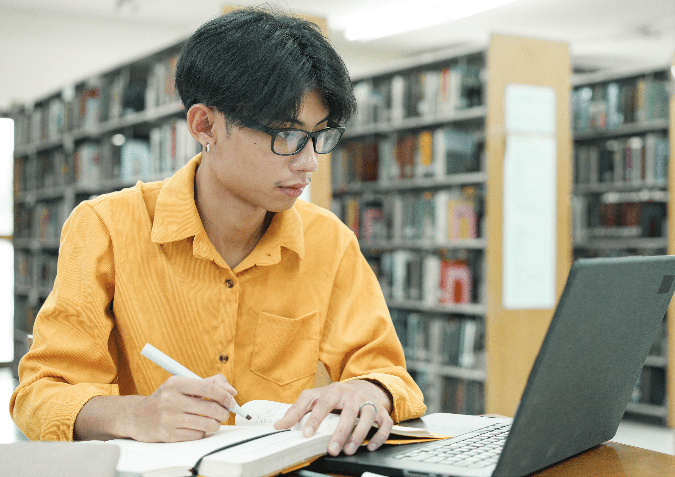 Young student wearing a yellow shirt studying with a laptop and notebook in a library, representing learning and professional development.