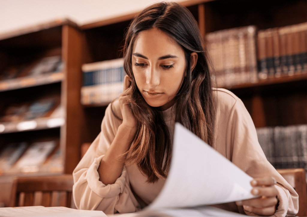 A young woman sitting in a library, reading documents with a focused expression. Bookshelves line the background, creating a quiet study atmosphere.