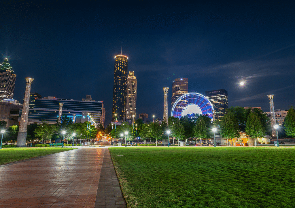 Night view of Centennial Olympic Park in Atlanta with illuminated skyscrapers, bright lights and a large circular attraction glowing against the dark sky.