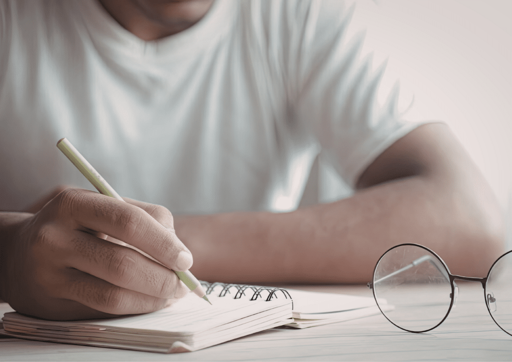 Close up of a person writing in a notebook with a pencil, glasses placed beside the notebook.